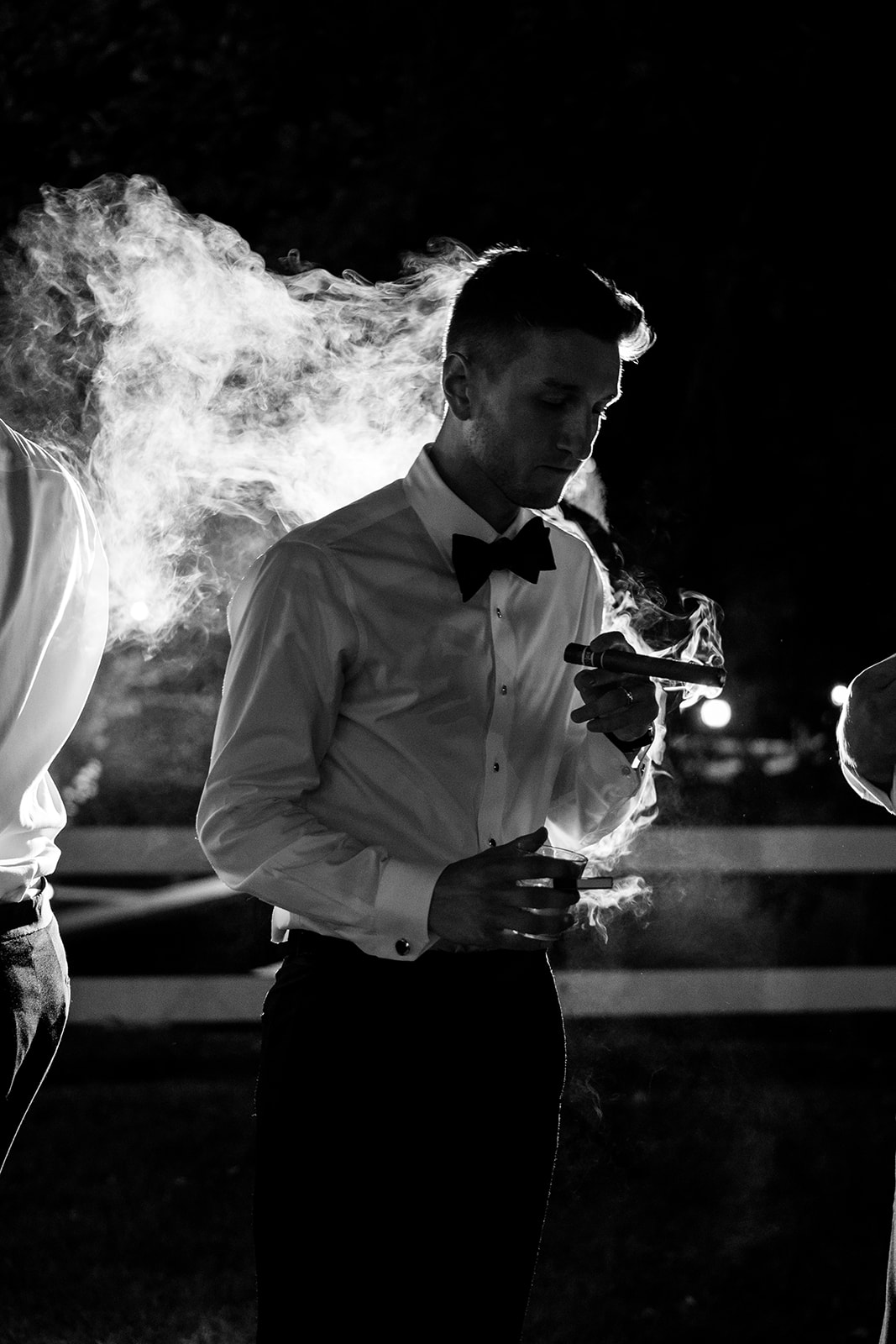 Black and white photo of the groom holding a cigar and a drink, backlit by glowing smoke at night.
